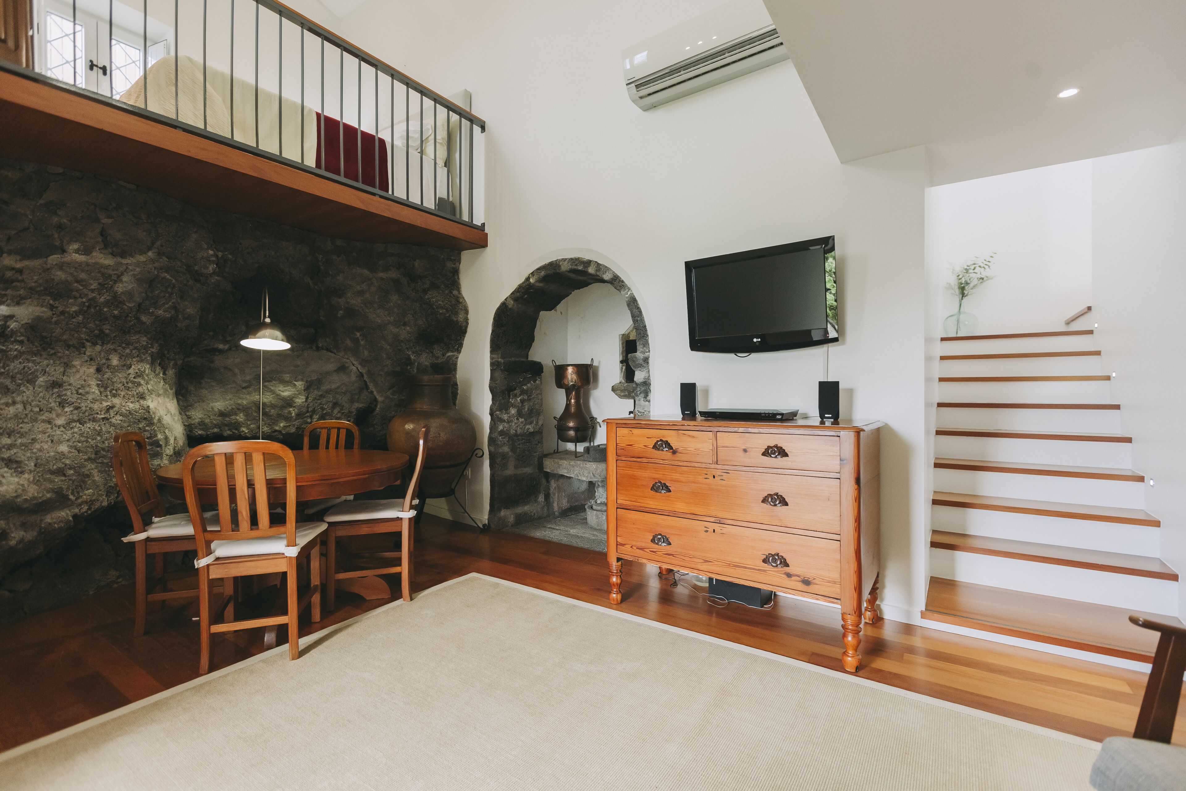 A dining area with a wooden table and chairs, a stone wall, a fireplace, and a staircase with a TV on the wall.