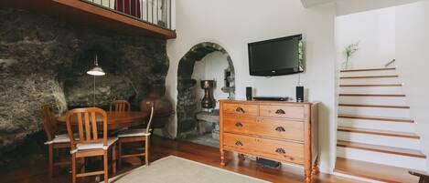 A dining area with a wooden table and chairs, a stone wall, a fireplace, and a staircase with a TV on the wall.