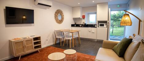 A modern living room with a sofa, two round tables, a dining area with chairs, a kitchen with a microwave, and a large window with a view of greenery.