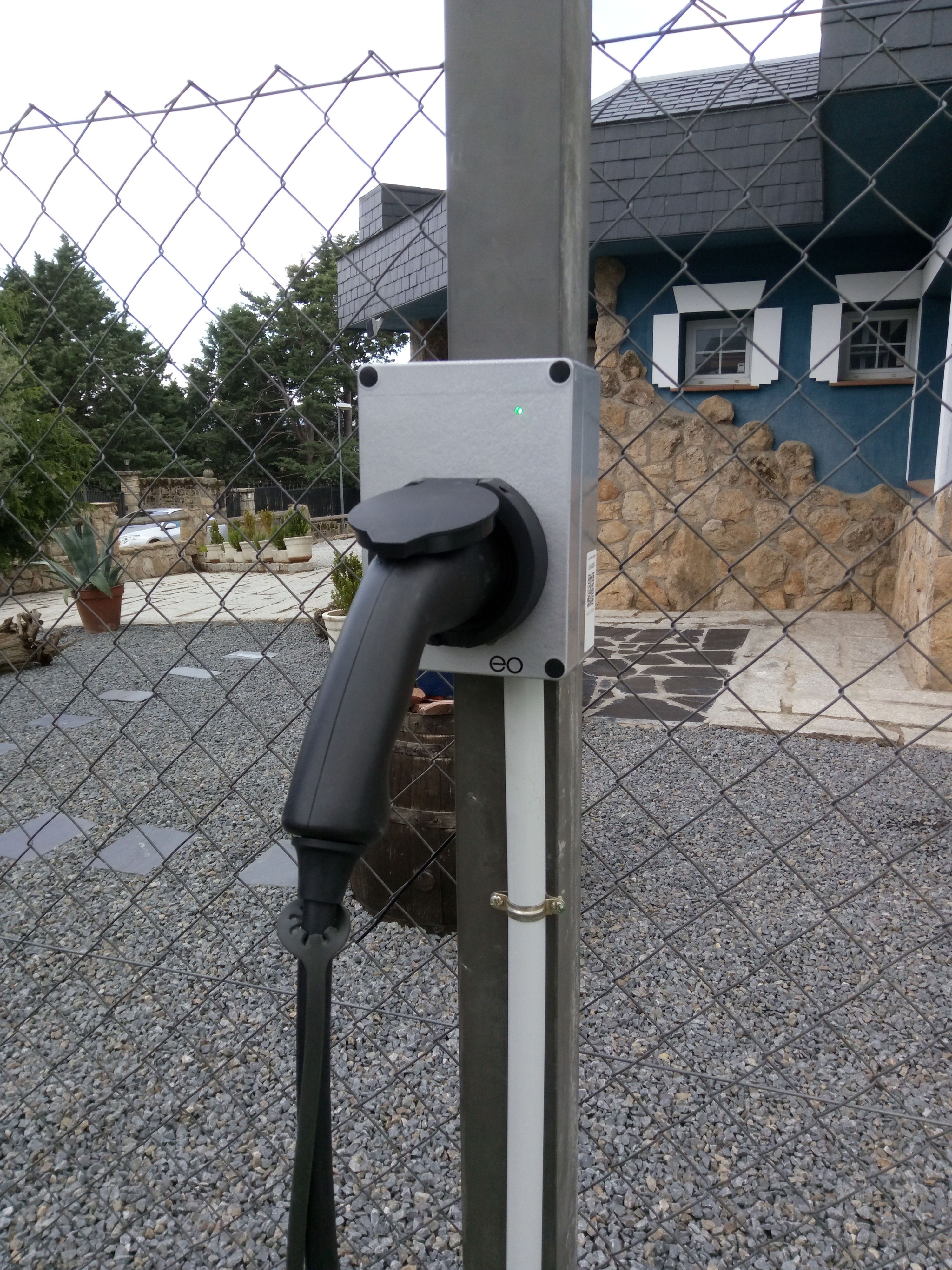 A parking meter with a charging station, a stone wall, and a house in the background.