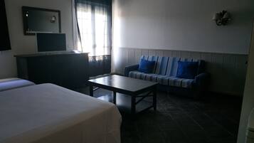 Habitación de hotel con dos camas, una mesita, un sofá con cojines azules, un televisor y una ventana con cortinas.