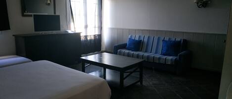 Hotellihuone, jossa on kaksi sänkyä, pieni pöytä, sinisillä koristetyynyillä varustettu sohva, televisio ja ikkuna, jossa on verhot.