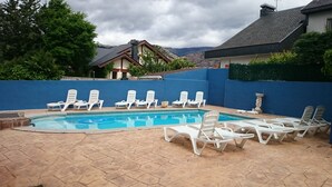 A swimming pool with white lounge chairs, a blue wall, and a house in the background. - Alojamiento la Pedriza (Manzanares el Real)