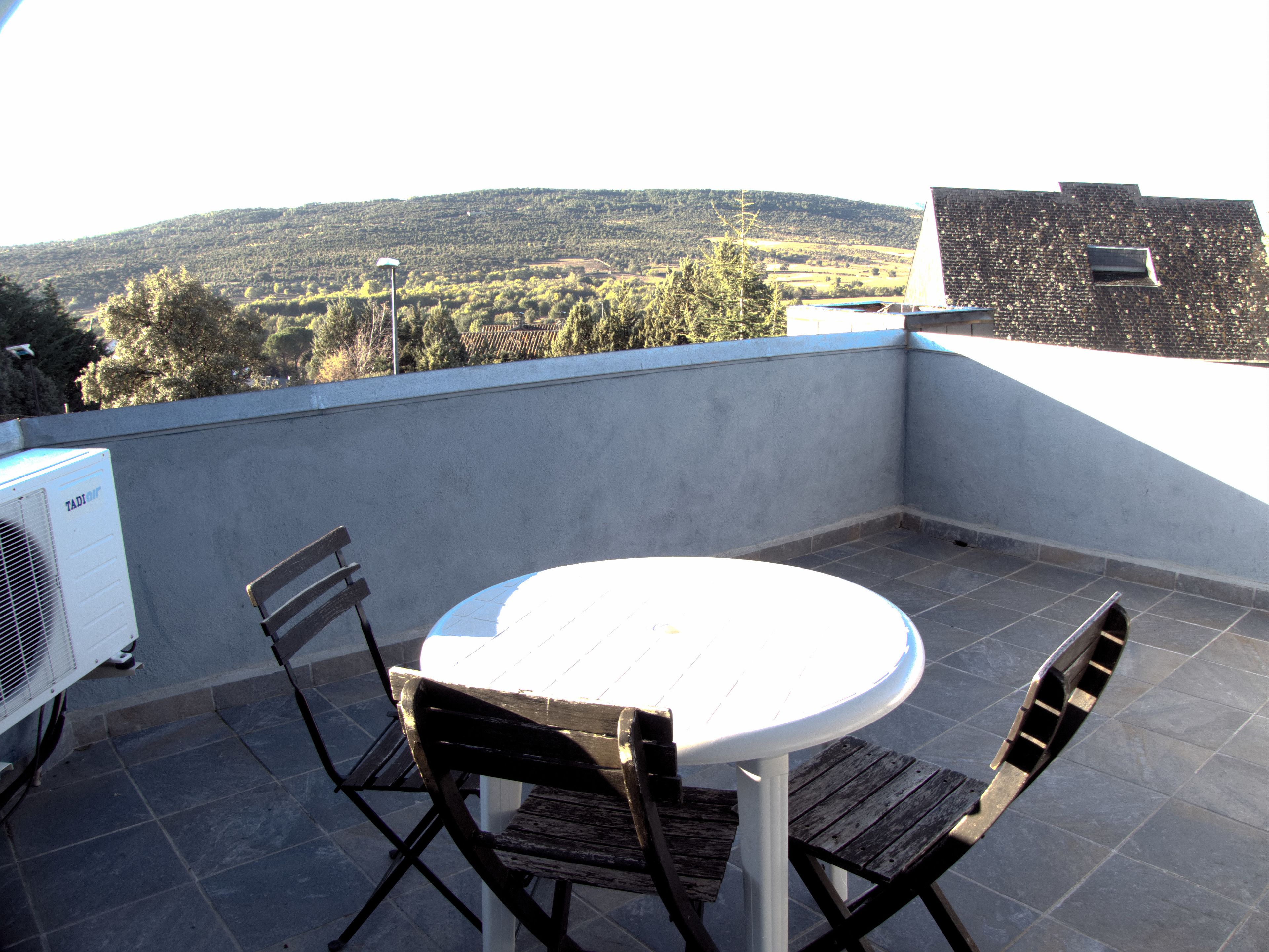 Un patio al aire libre con una mesa redonda y dos sillas, un equipo de aire acondicionado blanco y vistas a un paisaje montañoso.