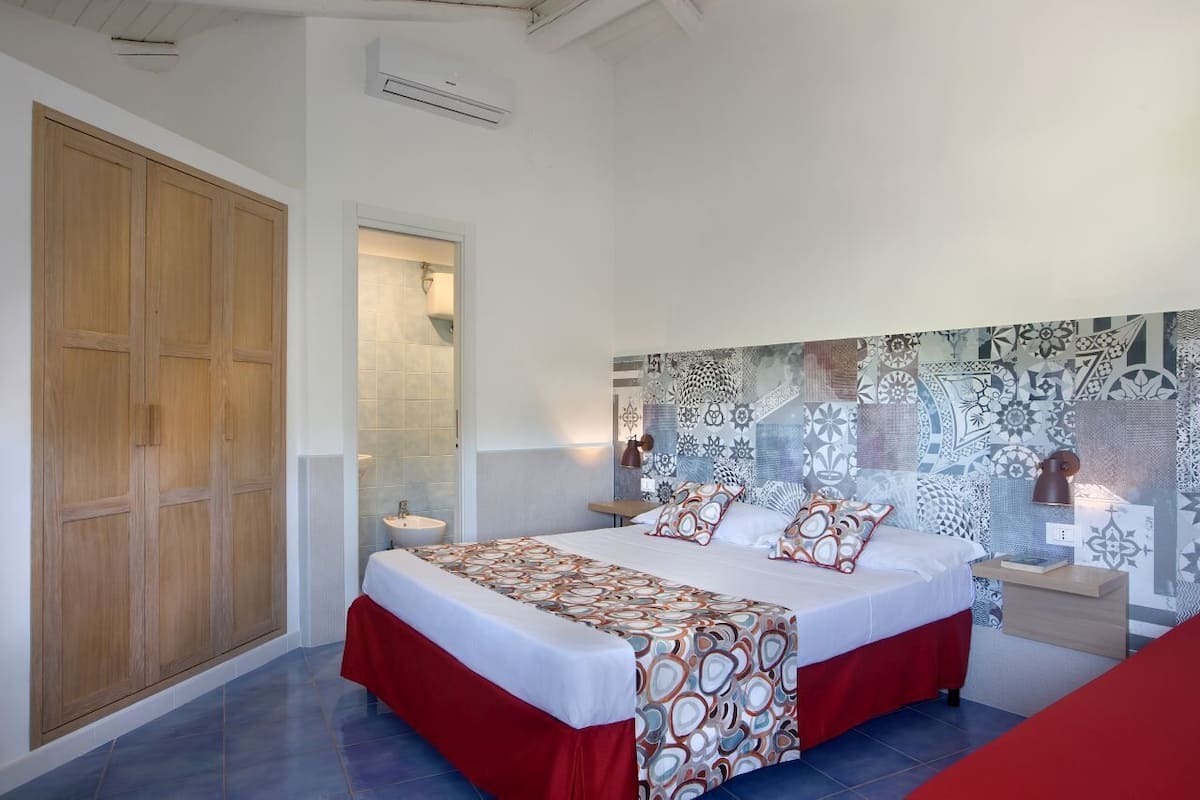 A bedroom with a large bed, a wooden wardrobe, a bathroom with a glass door, and patterned wall tiles.