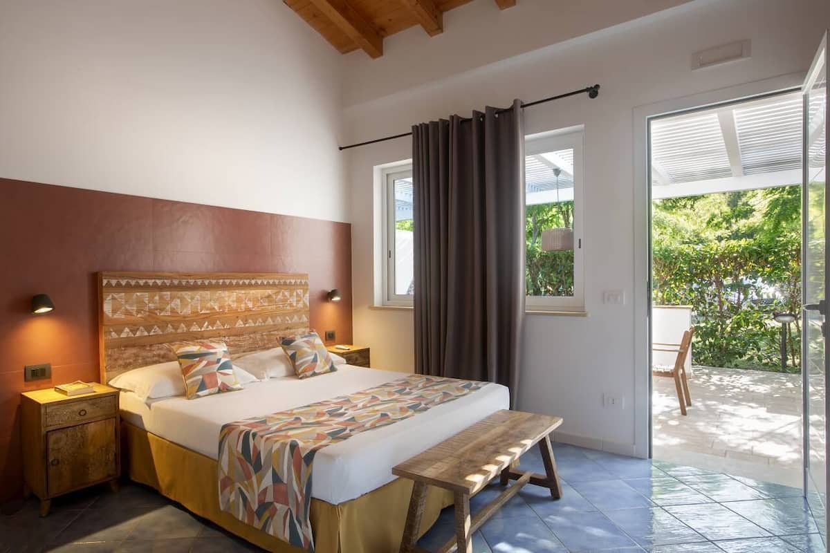 A bedroom with a large bed, a wooden bench, a nightstand, and a view of the garden through the window.