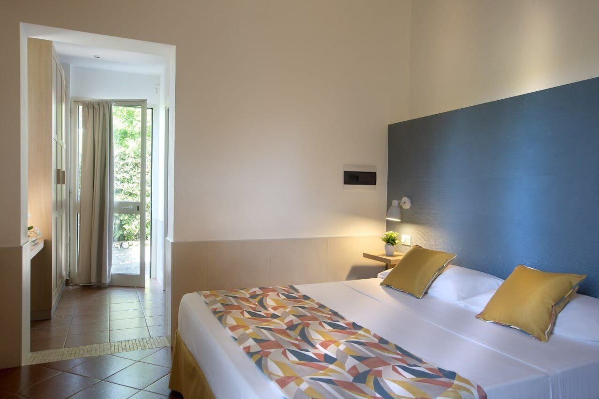A hotel room with a bed, a nightstand, a wall-mounted light, and a view of the outdoors through a door.