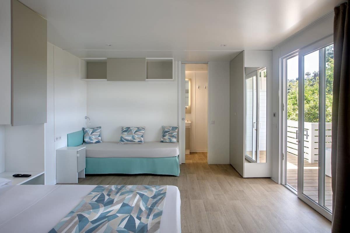 A modern bedroom with a bed, a sofa, and a small table. There is a sliding glass door leading to an outdoor area.