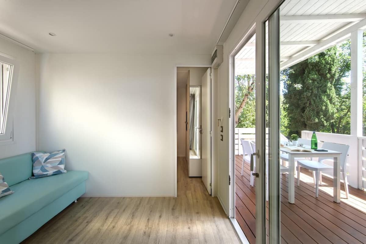 A modern interior with a teal sofa, a wooden floor, and a glass door leading to an outdoor deck with a table and chairs.