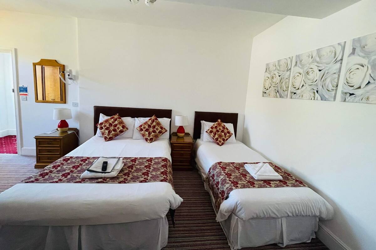 Une chambre d’hôtel avec deux lits, chacun recouvert d’une couvre-lit à motifs floraux et agrémenté d’oreillers assortis. On y trouve une table de chevet avec une lampe, une armoire en bois et un luminaire fixé au mur.