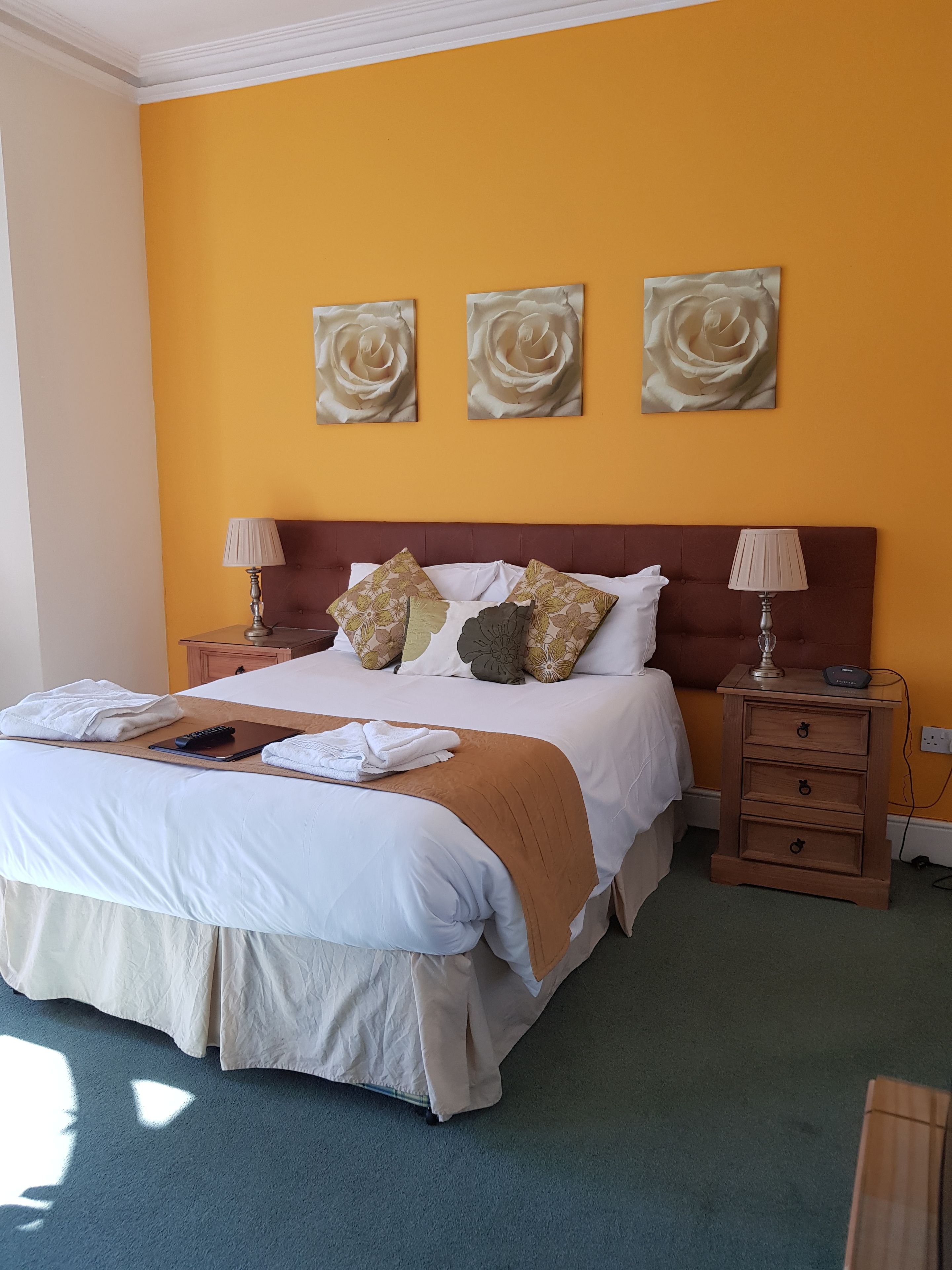A neatly made bed with a brown headboard, two bedside tables with lamps, and three wall-mounted rose paintings.