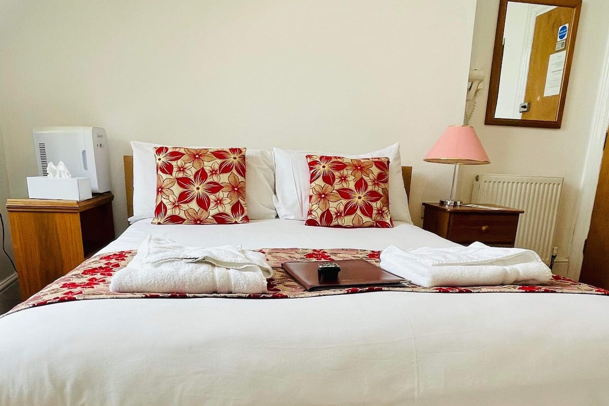 Un lit bien fait, avec du linge de lit blanc et des coussins à motifs floraux rouges, une table de chevet avec une lampe et un radiateur au mur.