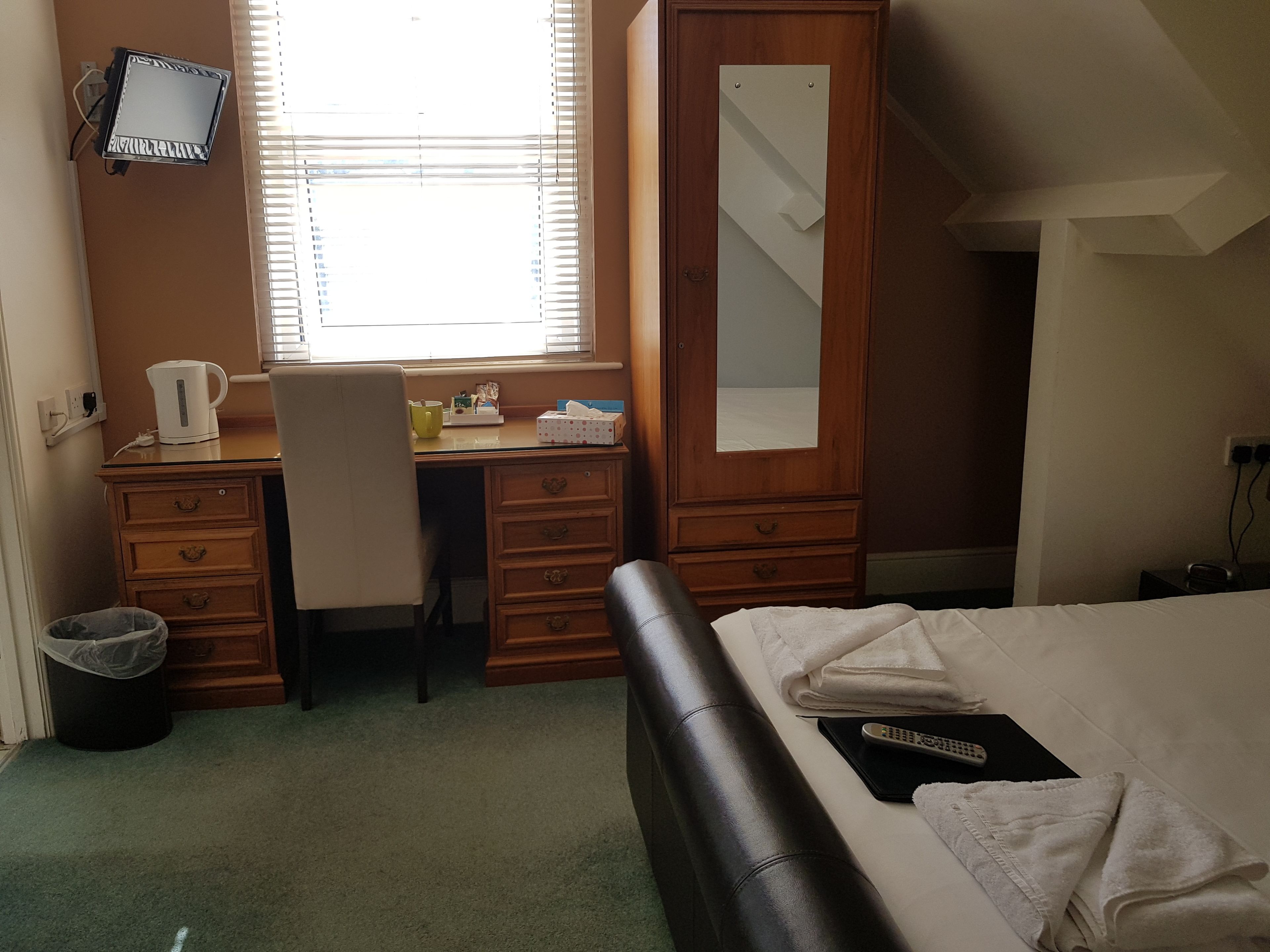 A hotel room with a bed, a desk with a chair, a wardrobe, a television, and a window with blinds.