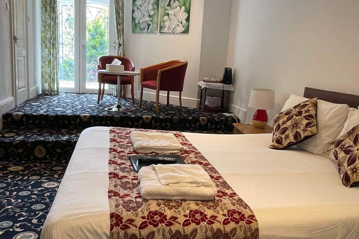 Une chambre d’hôtel avec un grand lit, un bureau et une chaise. Il y a une fenêtre avec des rideaux, un tableau au mur et une porte qui donne sur un balcon.
