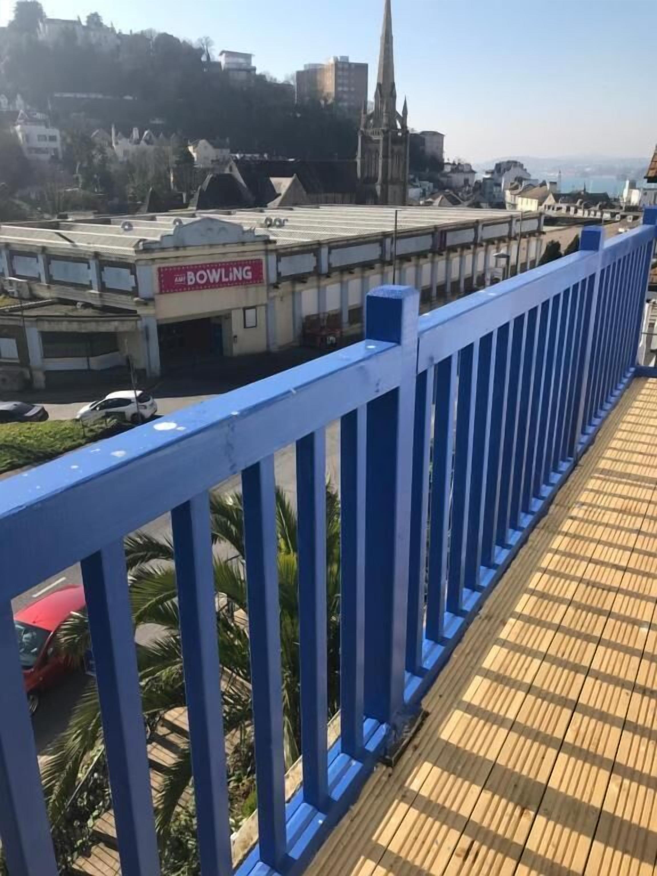 A wooden deck with a blue railing, overlooking a bowling alley and a church spire in the background.