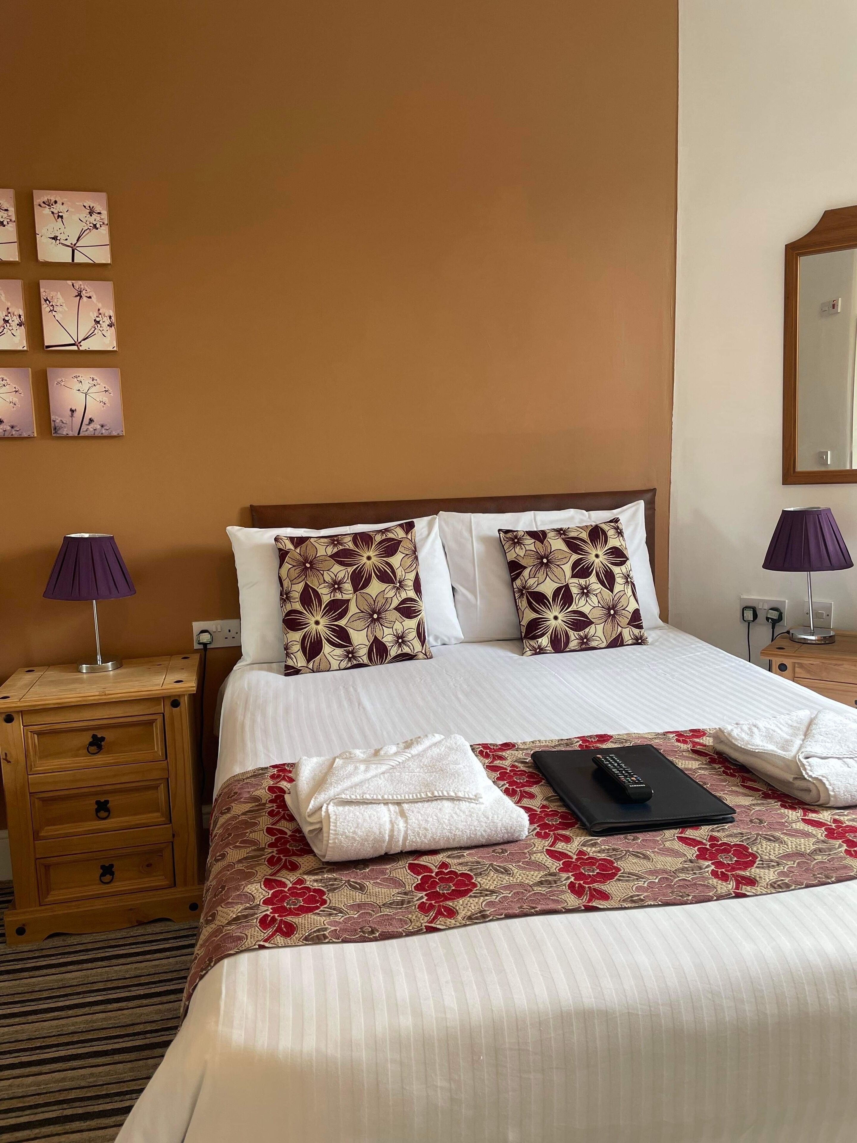 A neatly made bed with floral-patterned pillows, a bedside table with a lamp, and a wall with framed artwork.