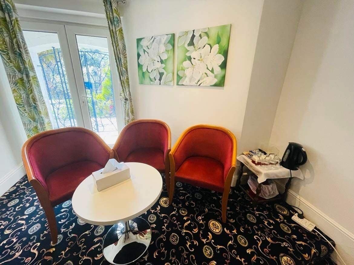 A room with two red armchairs, a small round table, and a window with curtains. There are two framed floral paintings on the wall.