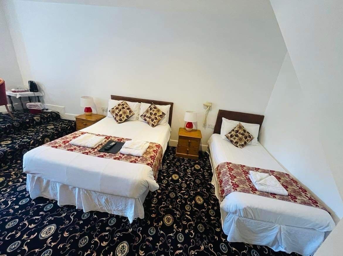 Une chambre d’hôtel avec deux lits, une moquette à motifs et une petite table avec une lampe.