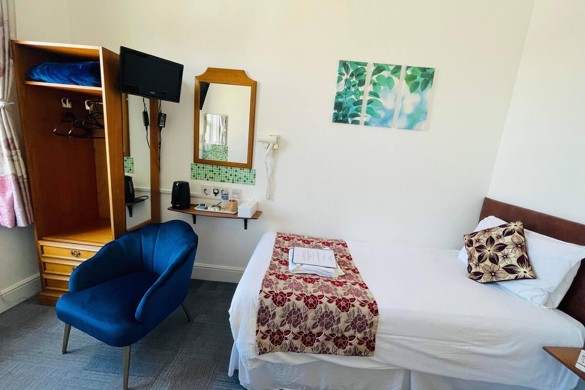 Une chambre d’hôtel avec un lit, une chaise bleue, un bureau avec un miroir, un téléphone et une petite étagère contenant des articles de toilette.