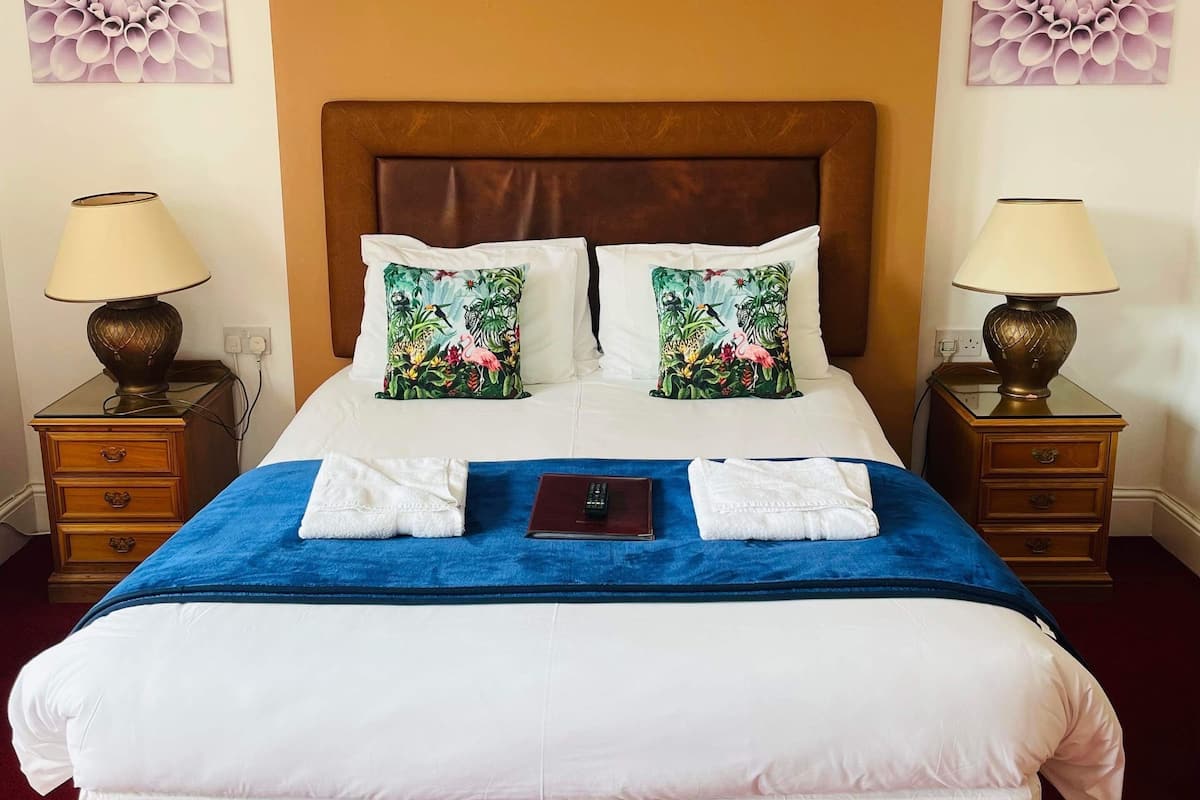 Un lit bien fait, avec du linge de lit blanc, deux coussins à motifs floraux, un plaid bleu et deux tables de chevet avec des lampes.