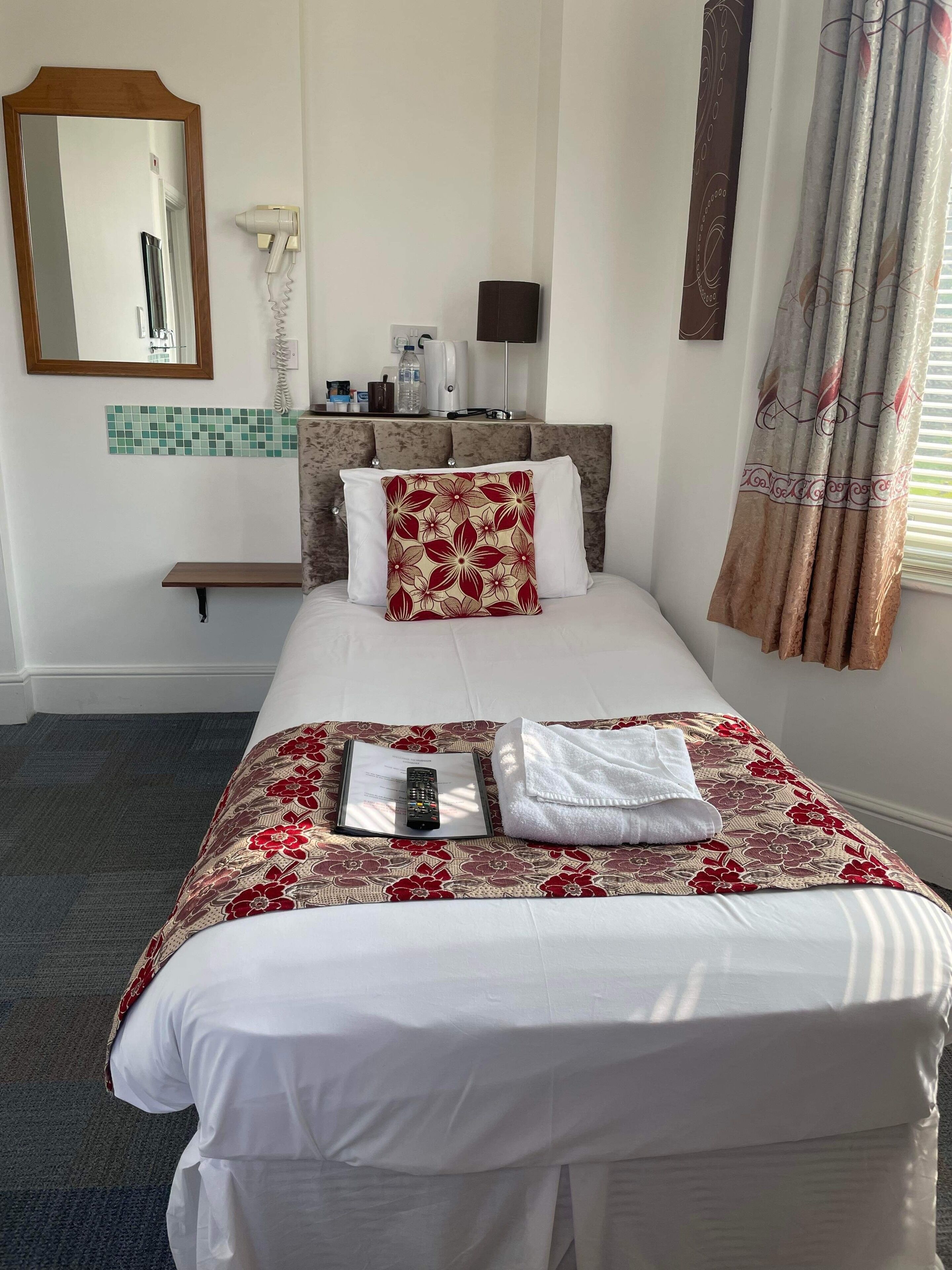 Une chambre d’hôtel avec un lit bien fait, une table de chevet avec une lampe, un miroir, un sèche-cheveux et une fenêtre avec des rideaux.