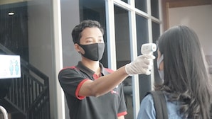Una persona con uniforme rojo y negro y máscara facial, sosteniendo un dispositivo para medición de temperatura.