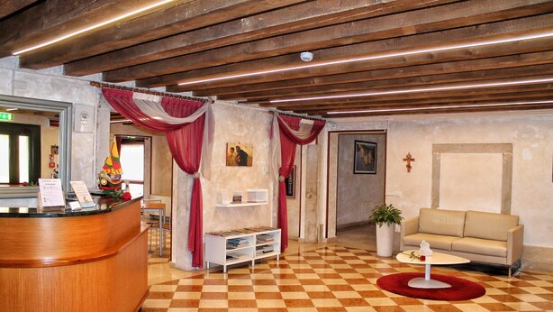 Ein Empfangsbereich mit Schachbrettmuster-Boden, einer Rezeption, einer Couch und einem Kruzifix an der Wand.