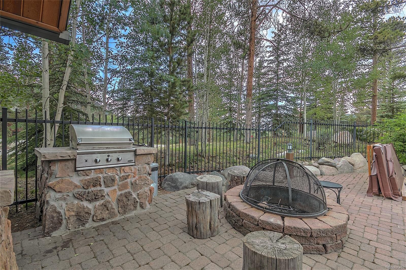 An outdoor patio with a stone barbecue grill, a fire pit, and a seating area surrounded by a metal fence and trees.