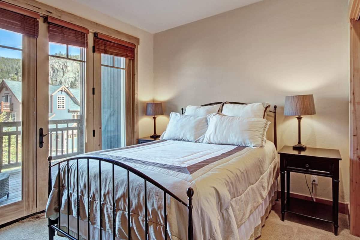 A bedroom with a bed, two bedside tables with lamps, a window with a view of a house, and a door leading to an outdoor area.