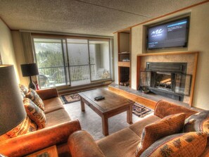 A living room with a fireplace, a television, and a large window with a view of trees. - Keystone Village Condos by Gondola Resorts (Keystone)