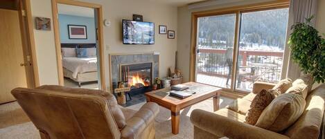 A cozy living room with a fireplace, comfortable seating, and a large window offering a view of a snowy landscape.
