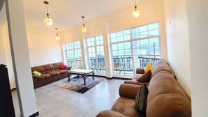 A spacious living room with a brown sofa, a coffee table, and large windows offering a view of a balcony. - Misthill Rest (Nuwara Eliya)