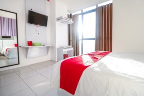 A hotel room with a bed, a TV mounted on the wall, a window with curtains, and a small shelf with a red cup and a green object. - RedDoorz near Terminal Batu Ampar (Balikpapan)