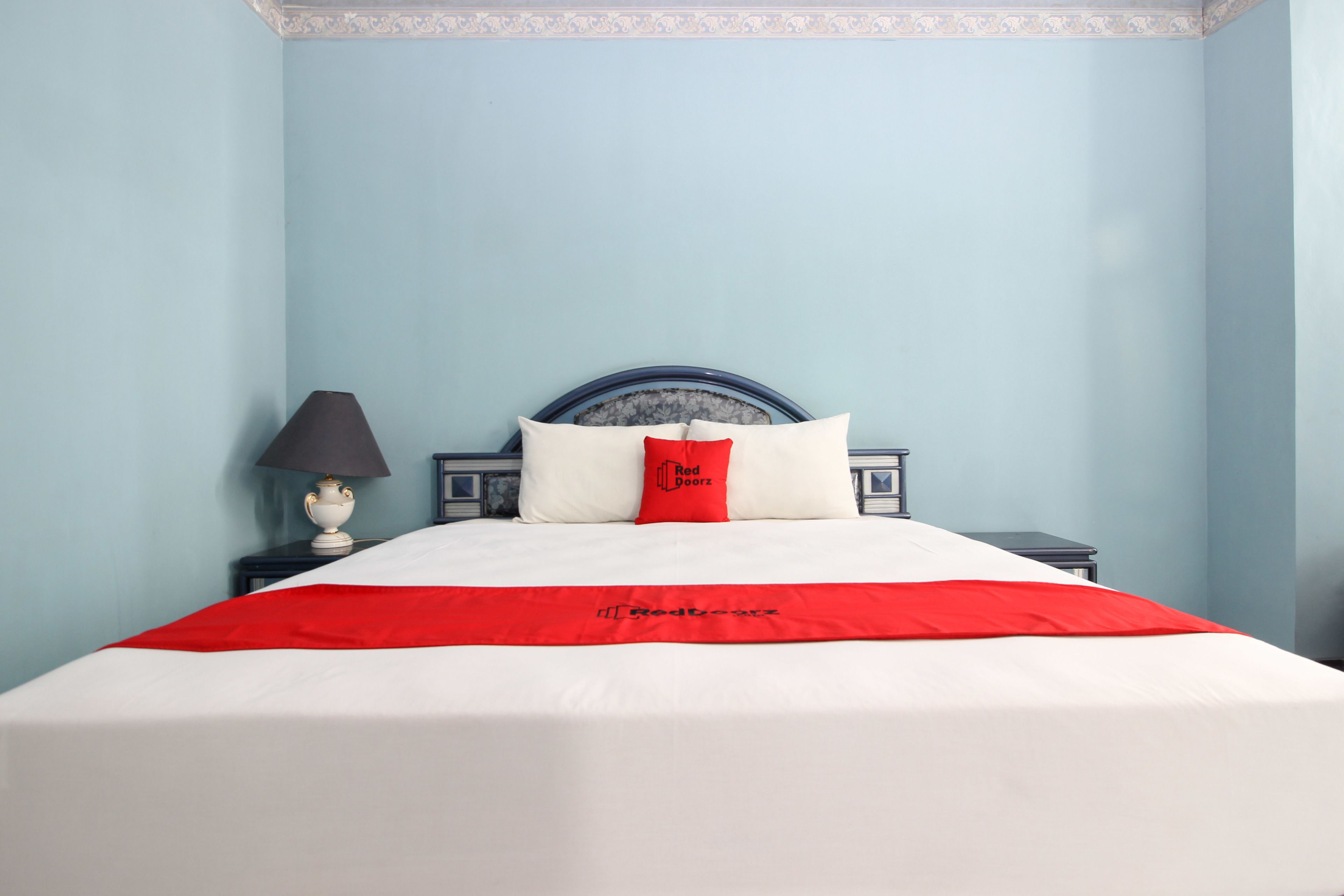 A neatly made bed with a white and red bedspread, a red pillow with "Red Doorz" text, and a black lamp on a bedside table.