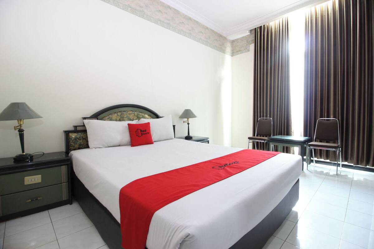 Quarto de hotel com uma cama grande, duas mesas de cabeceira com luminárias, uma pequena mesa com duas cadeiras e vista para o exterior através de cortinas.