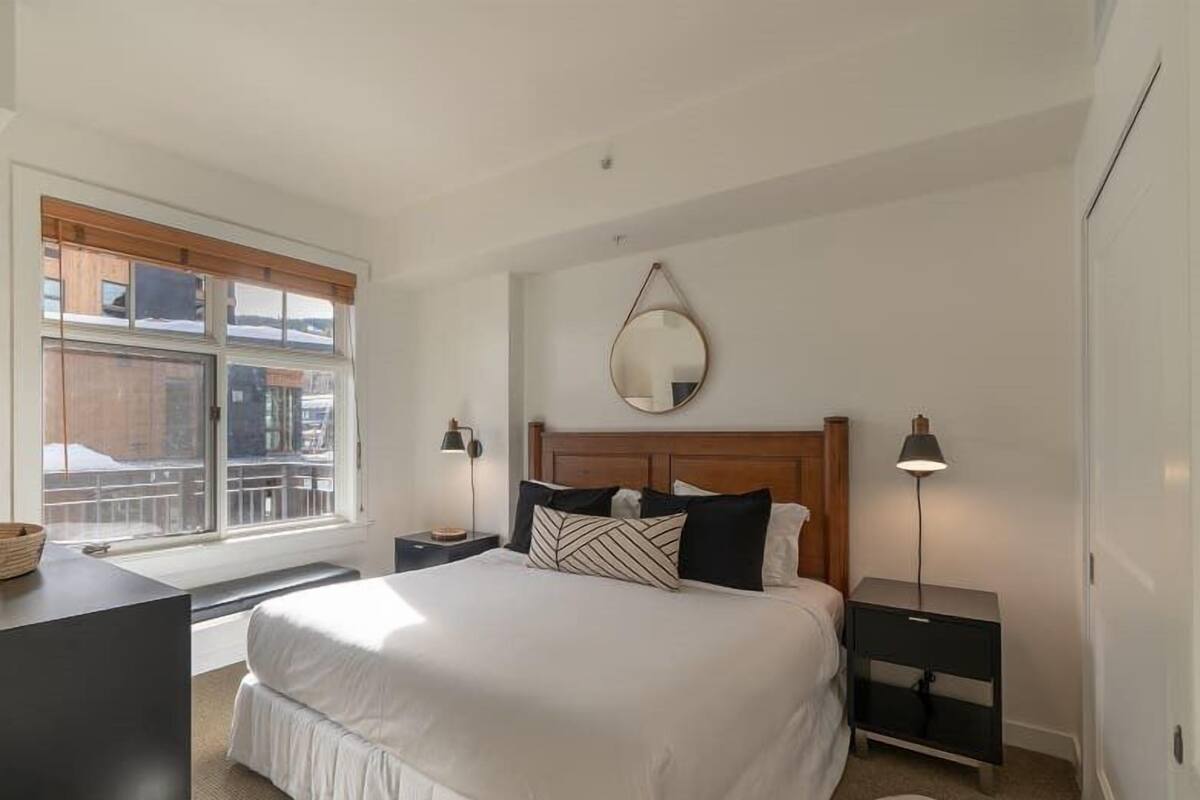 Un dormitorio con una cama grande, dos mesitas de noche, un espejo redondo y una ventana con vistas a edificios.