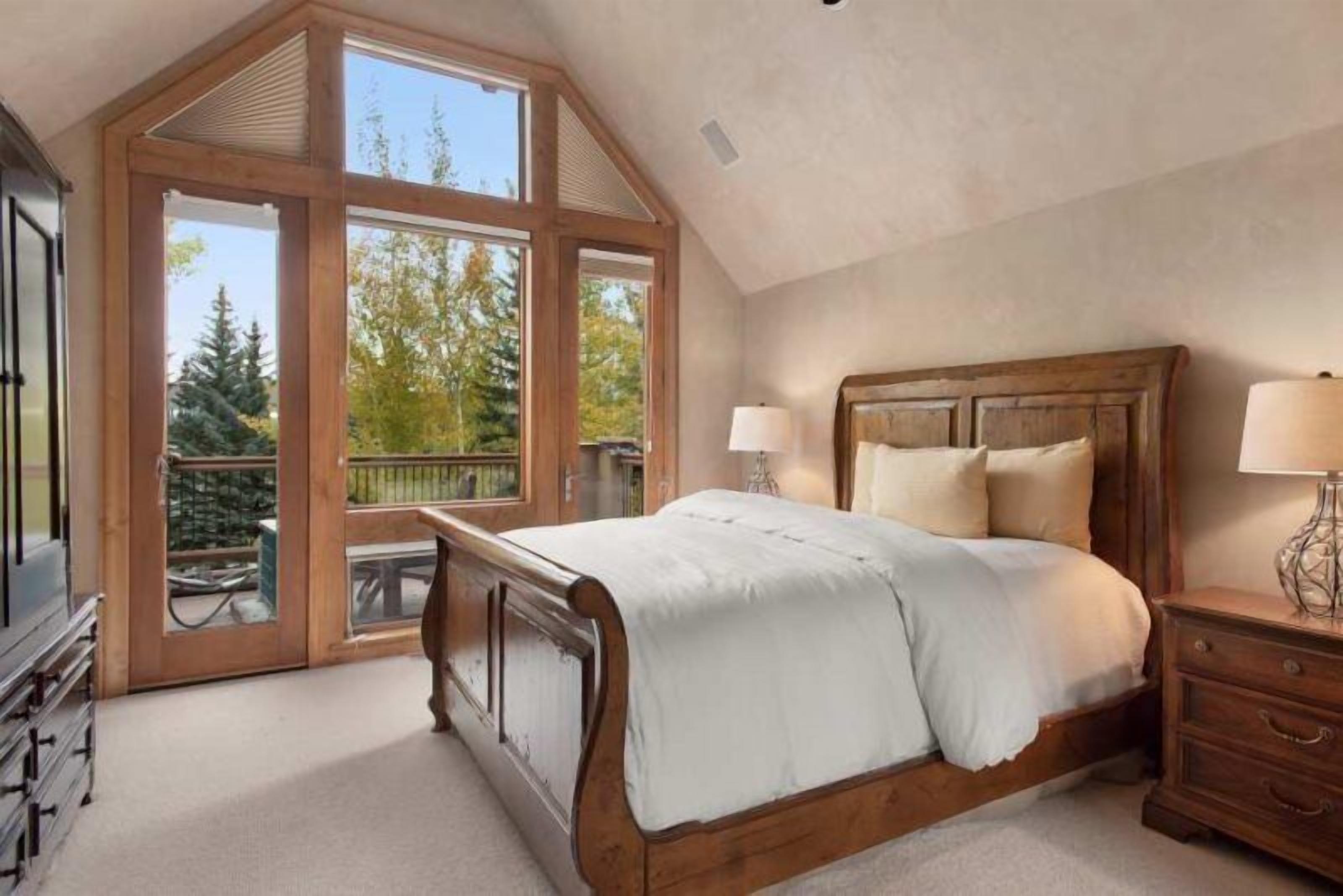 A bedroom with a wooden bed, two bedside lamps, and a large window with a view of trees.