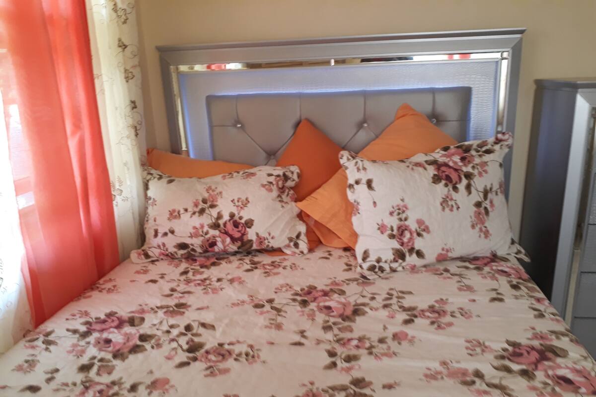 Una cama con cubrecama floral y dos almohadas naranjas, cabecera con diseño acolchado y una cortina roja visiblemente.
