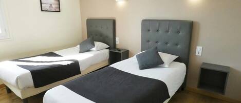Habitación de hotel con dos camas, cada una con cabecero gris, ropa de cama blanca y una manta color negro. Se observan mesitas de noche con lámparas, un aire acondicionado de pared y un cuadro enmarcado.