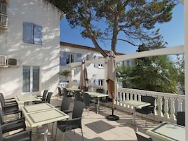 Un patio exterior con mesas y sillas, barandal blanco y un árbol que da sombra.