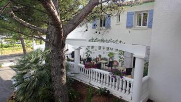 Un edificio blanco con balcón, mesas al aire libre y un árbol.