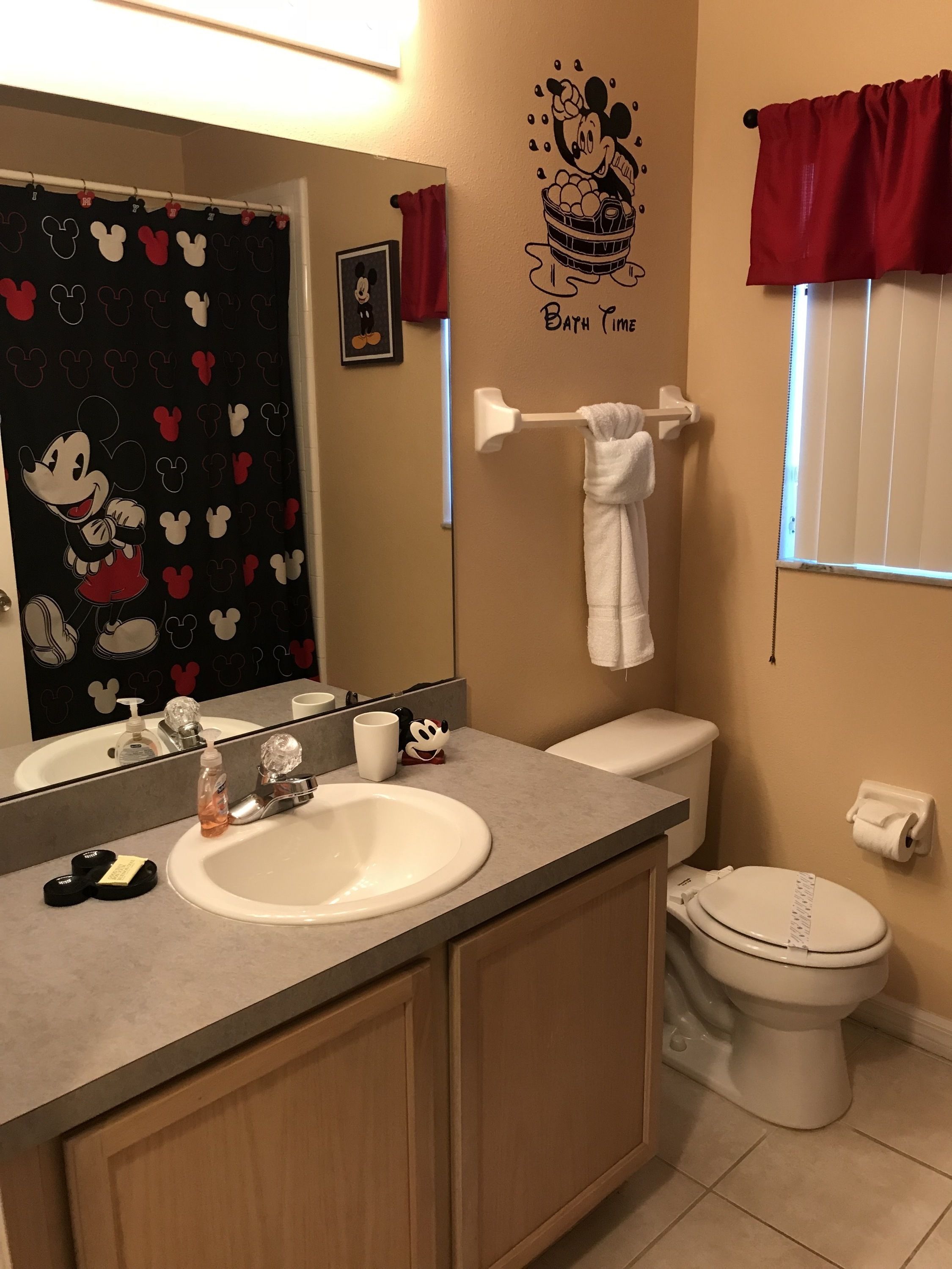 A bathroom with a Mickey Mouse themed shower curtain, a Mickey Mouse wall decal, a white sink, a toilet, and a towel rack.