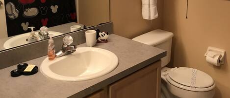 A bathroom with a Mickey Mouse themed shower curtain, a Mickey Mouse wall decal, a white sink, a toilet, and a towel rack.