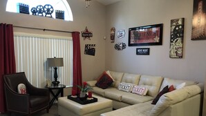 A living room with a sectional sofa, a small table, and a chair. There are movie posters and a clapperboard on the wall. - 4BR 3BA Home in Rolling Hiils by CV-7901 (Kissimmee)