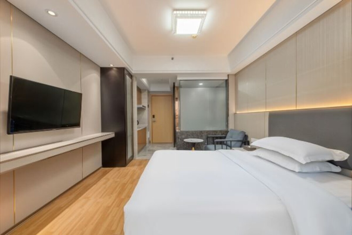 A modern hotel room with a large bed, a flat-screen TV mounted on the wall, a small table, and a seating area with a chair.