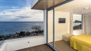 A balcony with a view of the sea, a bed with a yellow cover, and a small seating area. - Ocean Views by Azores Villas (Ponta Delgada)