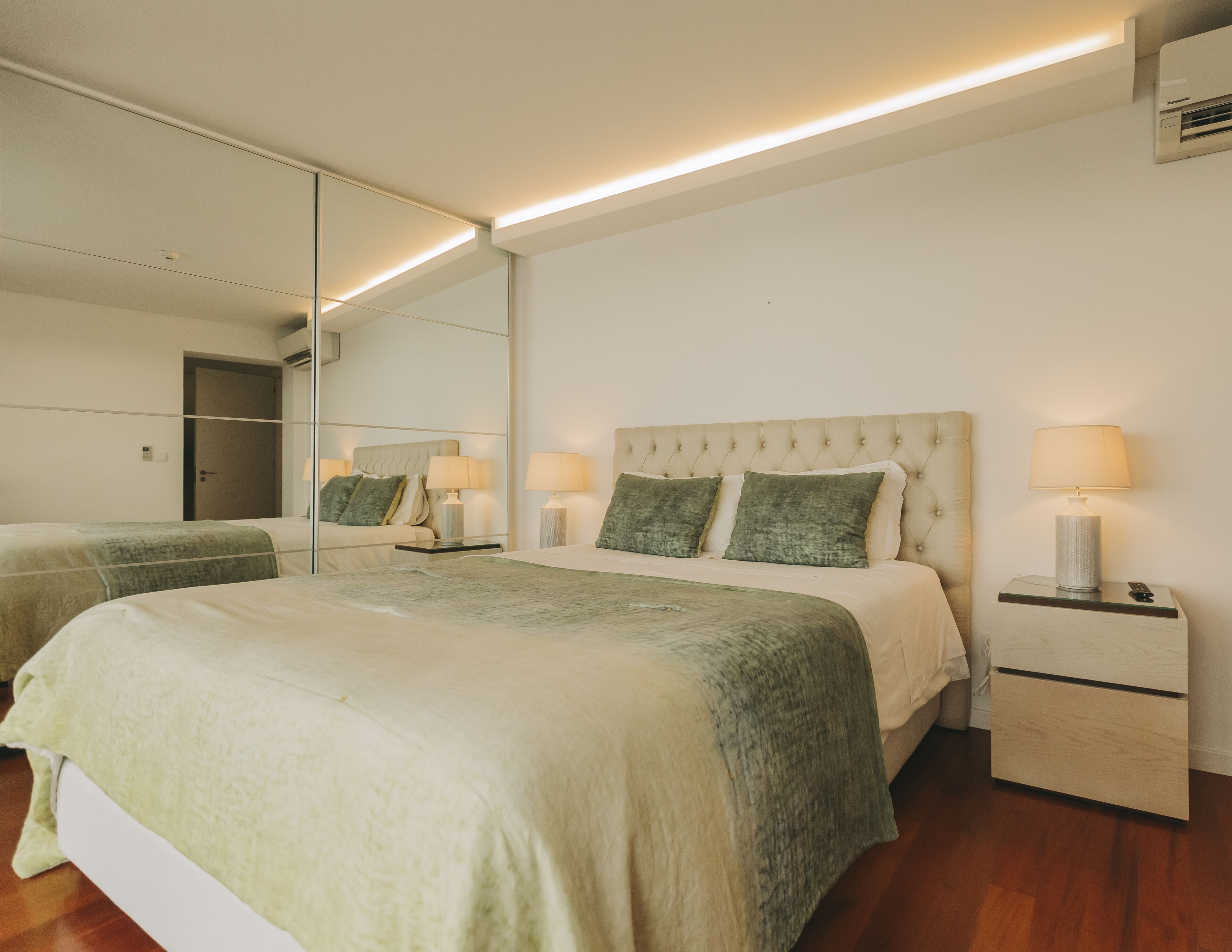 A modern bedroom with a large bed, two bedside tables, a mirror, and a wall-mounted air conditioner.