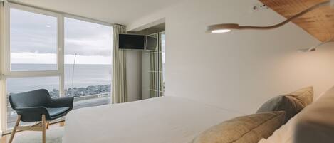 A modern hotel room with a large bed, a chair, a TV, and a view of the ocean.