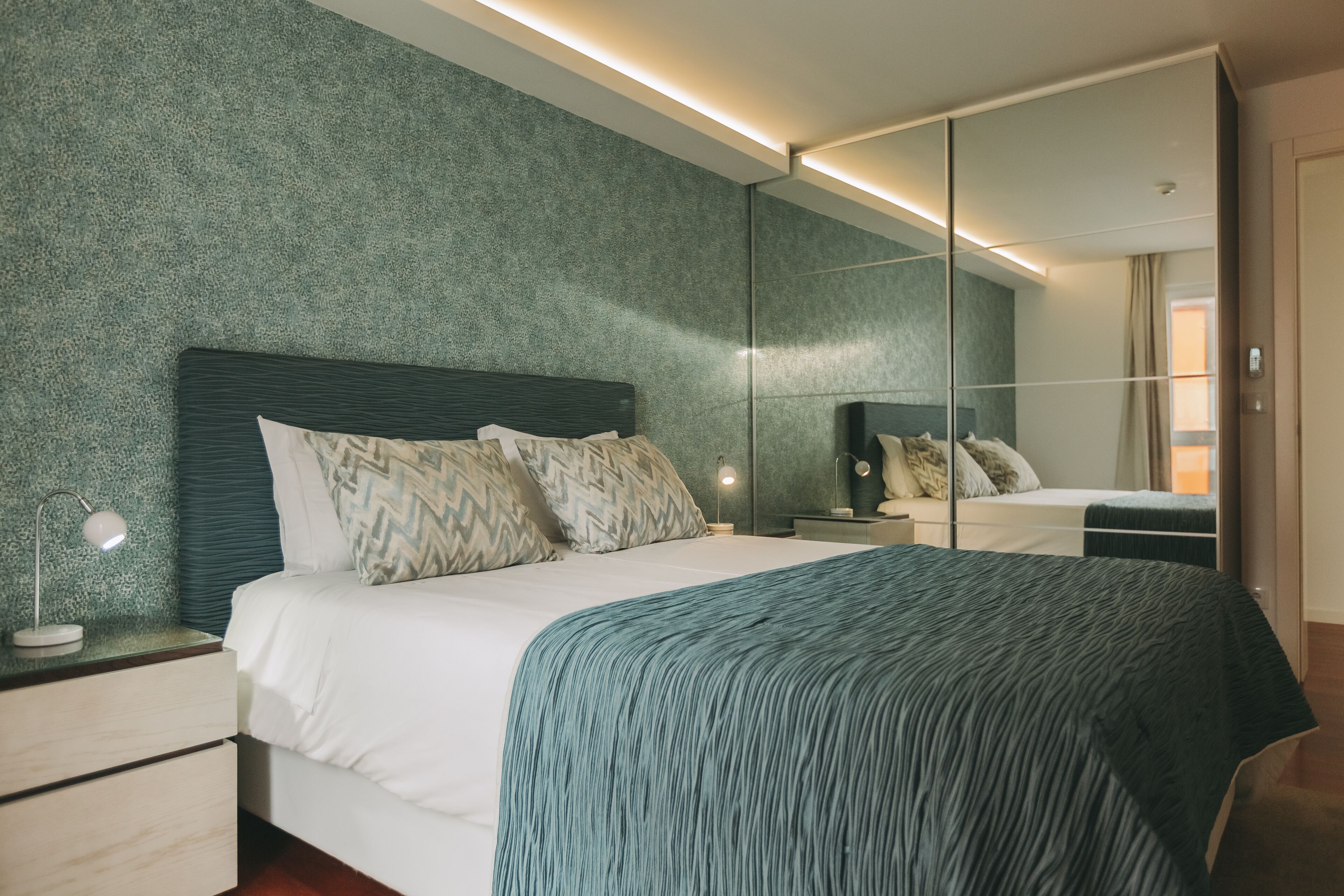 A modern bedroom with a large bed, a nightstand with a lamp, a dresser, and a mirror.