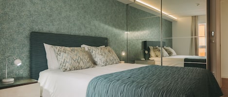 A modern bedroom with a large bed, a nightstand with a lamp, a dresser, and a mirror.